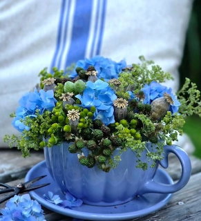 Blue ceramic teacup filled with hydrangeas, green berries, and wildflowers on a rustic wooden surface