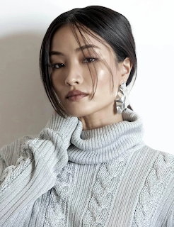 Woman in white cable-knit turtleneck sweater with dark hair styled back, wearing statement earrings