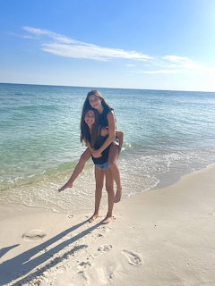 Two girls standing on a sandy beach by the ocean, one giving the other a piggyback ride.