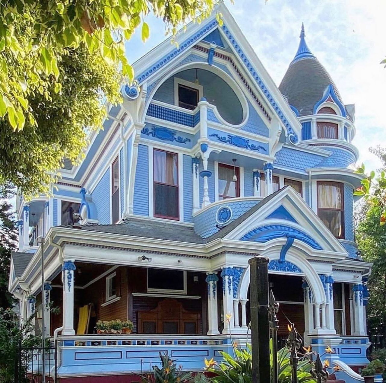 Ornate blue Victorian house with white trim, turret, and wraparound porch surrounded by greenery