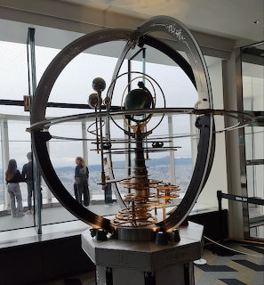 Mechanical orrery solar system model in a modern glass observation deck with city views behind