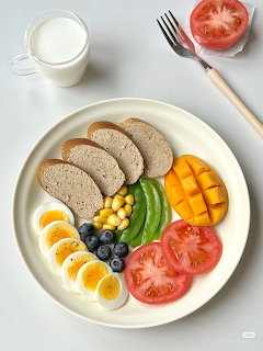 White plate with bread slices, boiled eggs, tomatoes, mango, avocado, blueberies, corn, glass of milk and fork