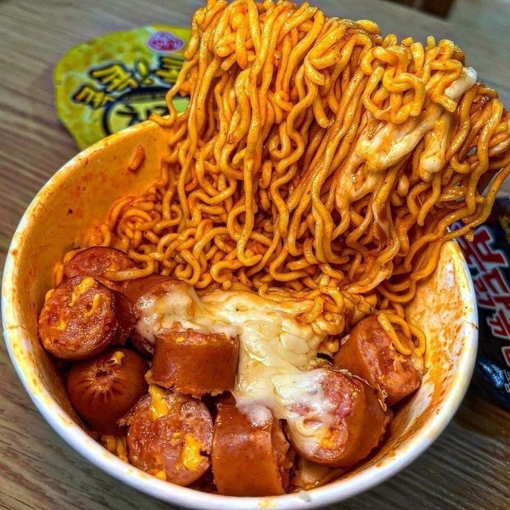 Bowl of spicy ramen with sliced sausage and melted cheese on a wooden table