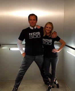 Man and woman posing together wearing matching black NCIS 300 t-shirts indoors