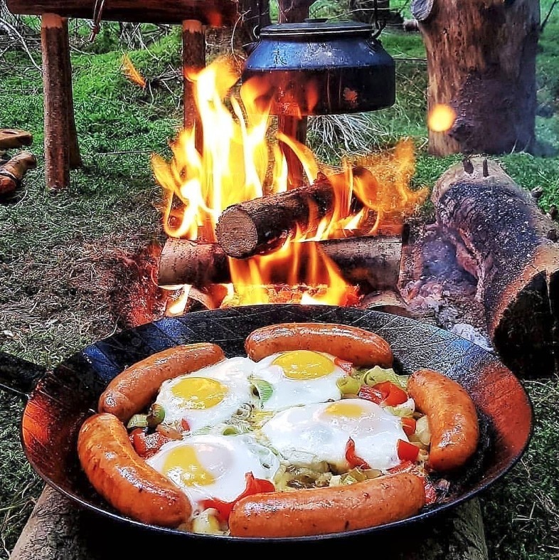 Sausages and fried eggs cooking in skillet over campfire with flames and hanging kettle
