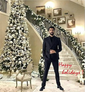 Man in black suit standing next to a white Christmas tree in a grand staircase interior