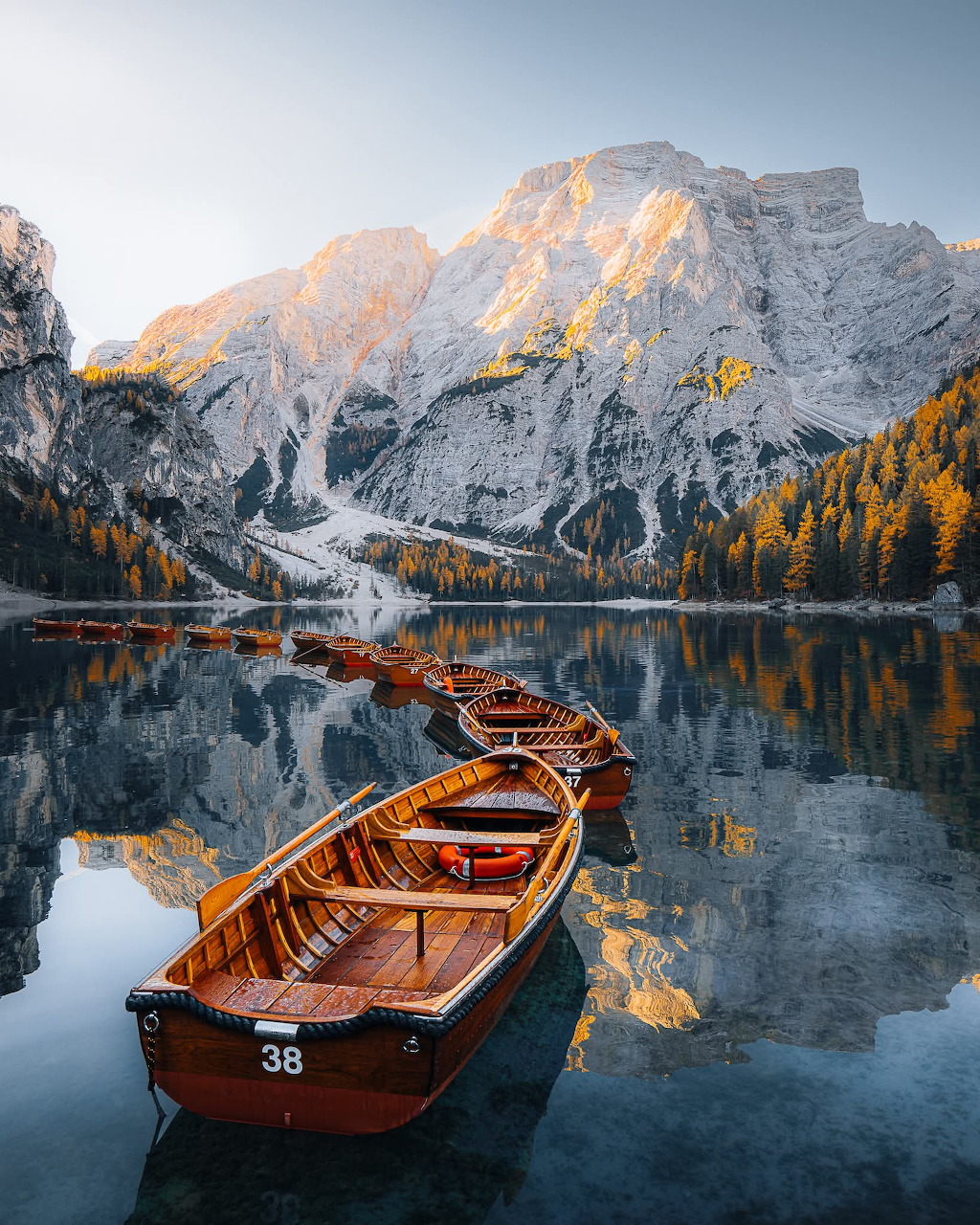 Wooden rowboats on a tranquil alpine lake with autumn trees and snowy Dolomite peaks reflected in the water.
