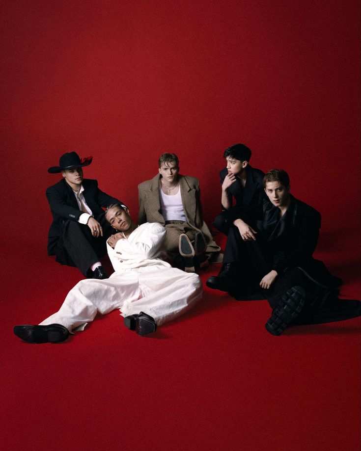 Five men in formal black and white clothing pose together on a red background