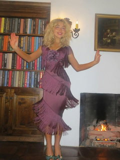 Blonde woman in a fringed purple dress posing beside a bookshelf and fireplace indoors.