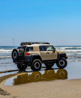 Tan Toyota FJ Cruiser with roof rack and off-road tires driving on a sandy beach near the ocean