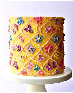 Yellow decorated cake with piped diamond pattern and colorful fondant flowers on a white stand