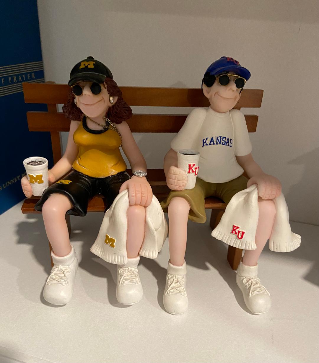 Two ceramic figurines of college sports fans sitting on a bench, one in Michigan gear and one in Kansas gear.