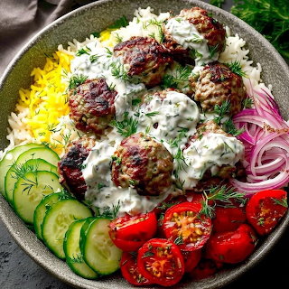 Bowl of meatballs with yogurt sauce on rice, garnished with cucumber slices, cherry tomatoes, and pickled red onions