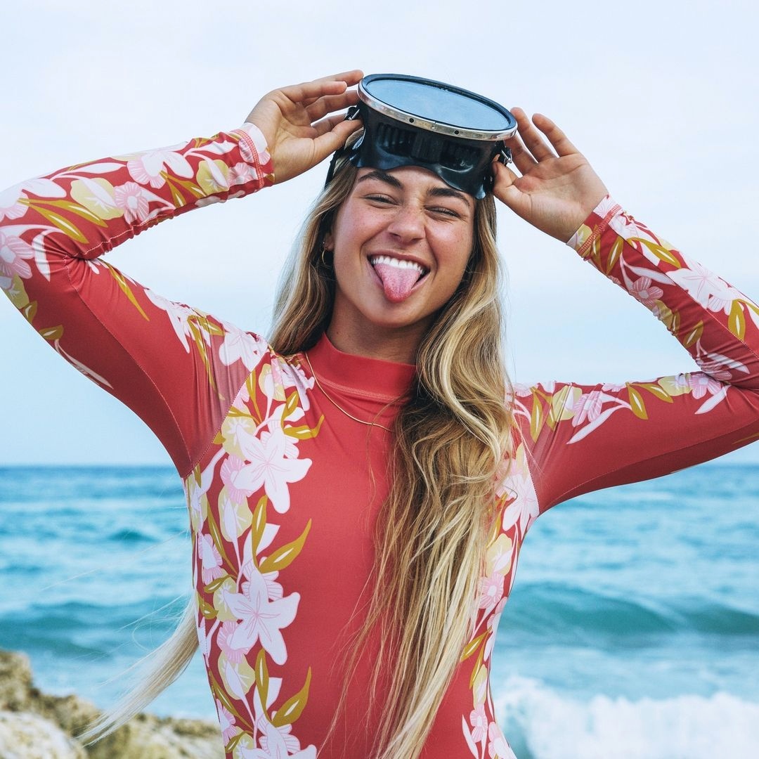 Blonde woman in red floral wetsuit holding goggles on head, sticking tongue out at beach