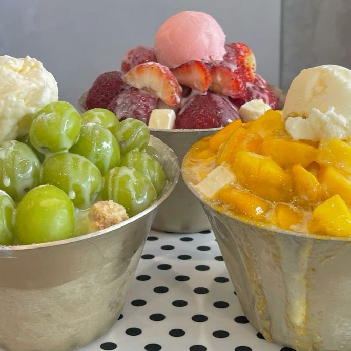Three shaved ice desserts with grapes, strawberries, mango, ice cream and whipped cream in bowls