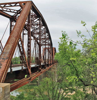 Rusty railroad bridge over the Colorado River with green trees and a cloudy sky
