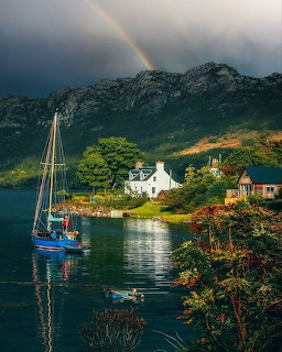 Scottish Highland village with white houses by loch, sailboat, mountains and rainbow