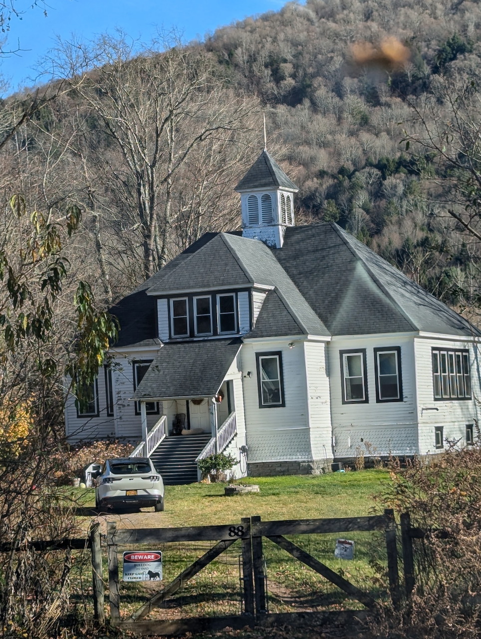 White church converted into a home with cupola, set against a wooded hillside with a wooden fence in front