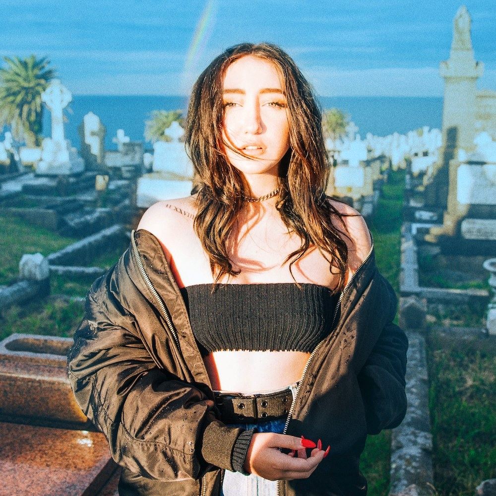 Young woman with long brown hair in black top outdoors with coastal cemetery background