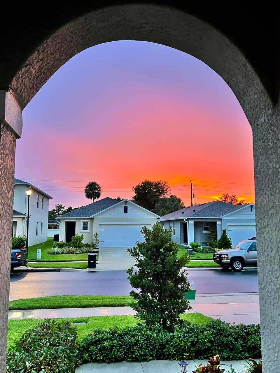 Colorful sunset viewed through stone arch framing suburban neighborhood with houses and green lawn