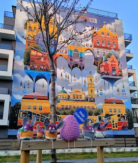 Colorful painted mural on building wall showing city landmarks with crocheted figures in foreground