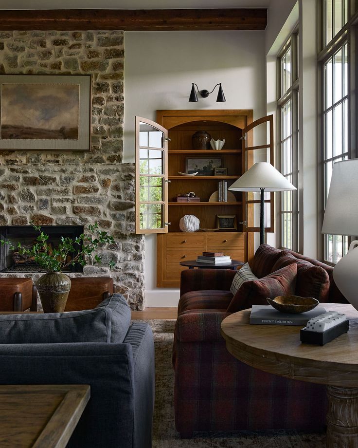 Rustic living room with stone fireplace, plaid sofa, large windows, and wooden shelves