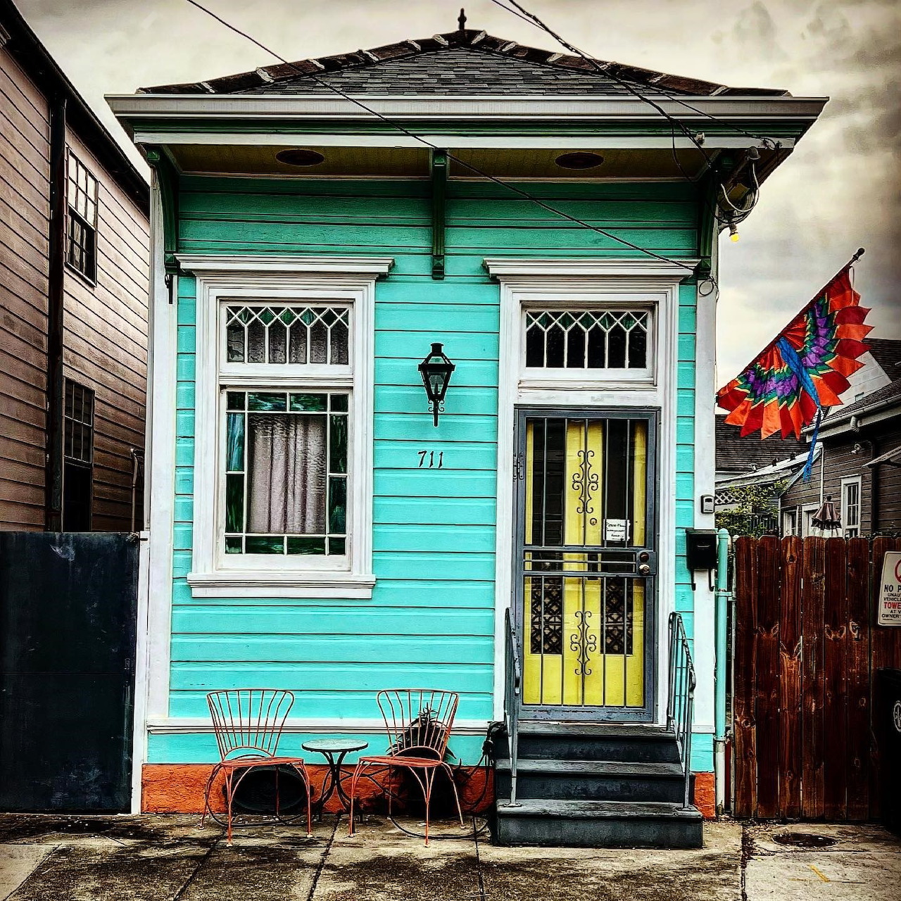 Small turquoise shotgun house with yellow door, number 711, and matching chairs on the sidewalk