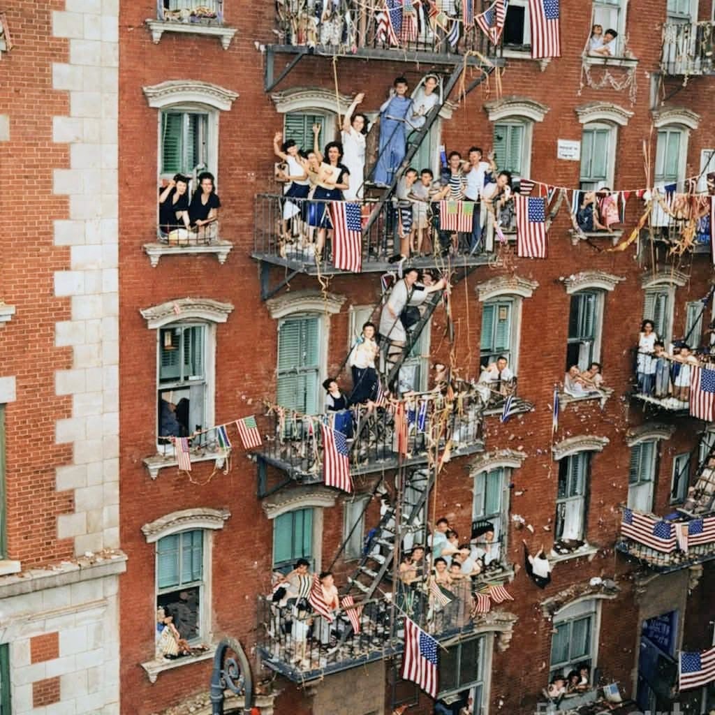 Vintage colorized photo of people on fire escapes and windows of a brick apartment building with American flags