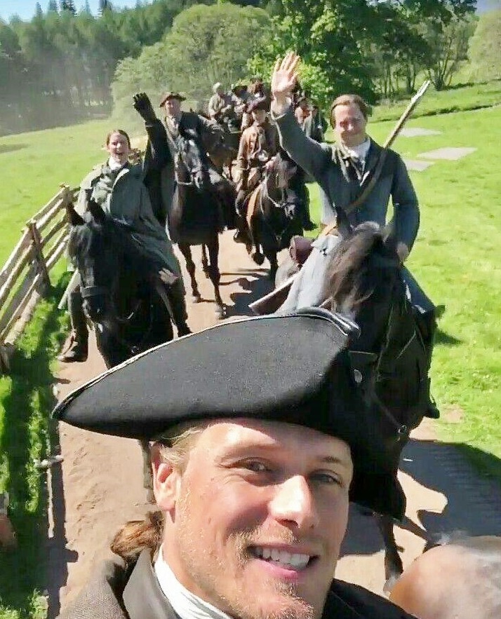Smiling person in tricorn hat taking selfie with costumed group on grassy hillside