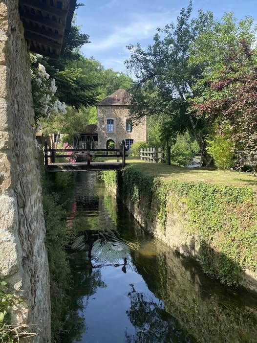 Narrow canal beside a stone wall leading to a small house and bridge among trees
