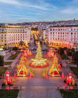 Festive city square with a lit Christmas tree, decorations, and surrounding buildings at dusk