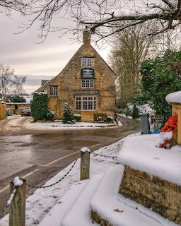 Snow-covered Cotswold stone pub with war memorial and bare trees in winter