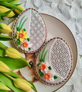 Two oval cookies decorated with lace icing patterns and floral designs, arranged with yellow tulips on white textured background
