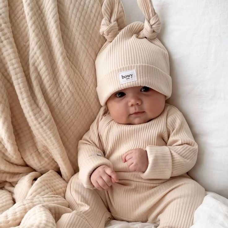 Baby in beige ribbed clothing and knotted hat lying on cream blankets