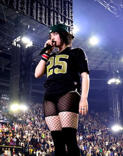 Female singer on stage with microphone wearing black jersey number 25, green beanie, and fishnet stockings
