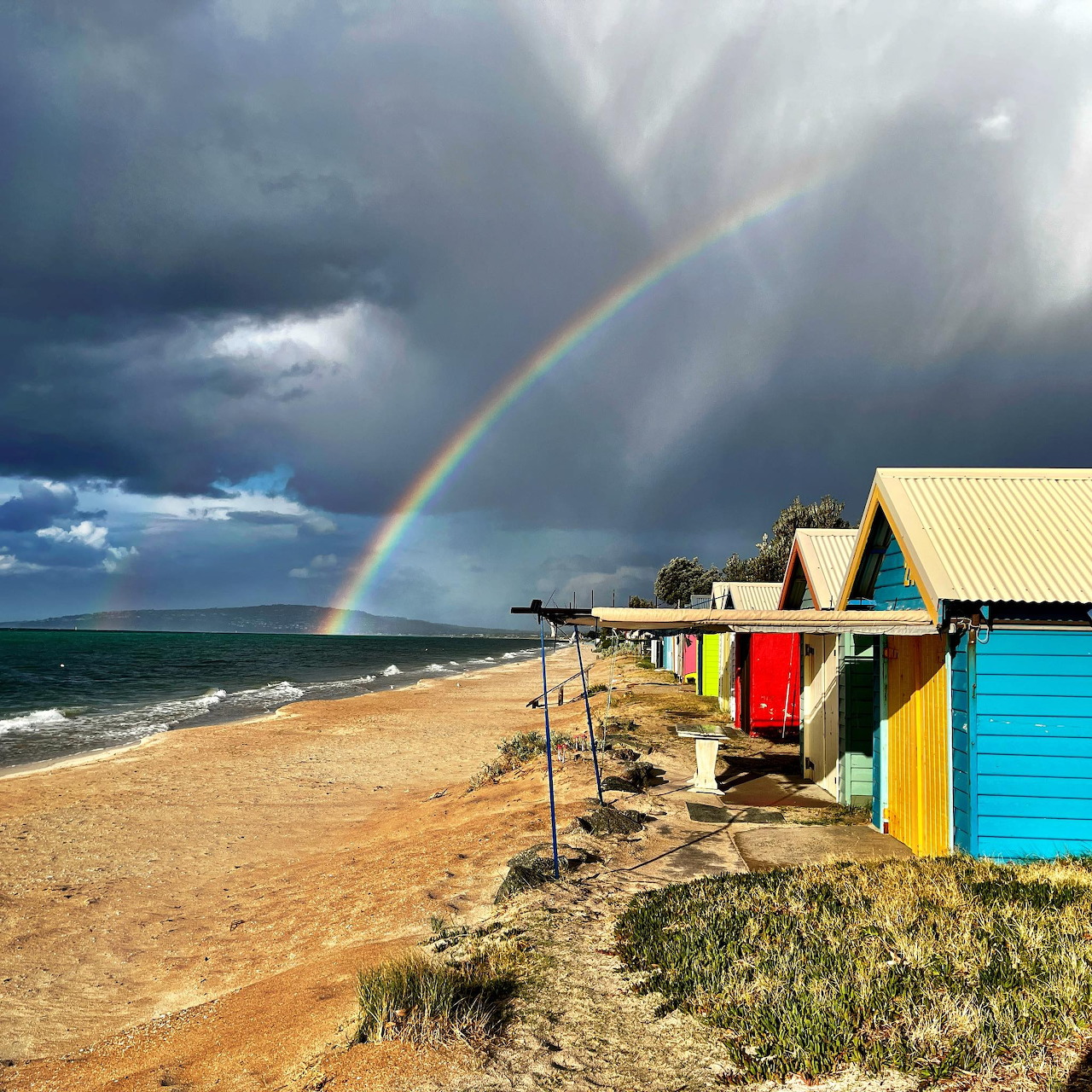 Rainbow over colorful beach huts on sandy shore with stormy clouds