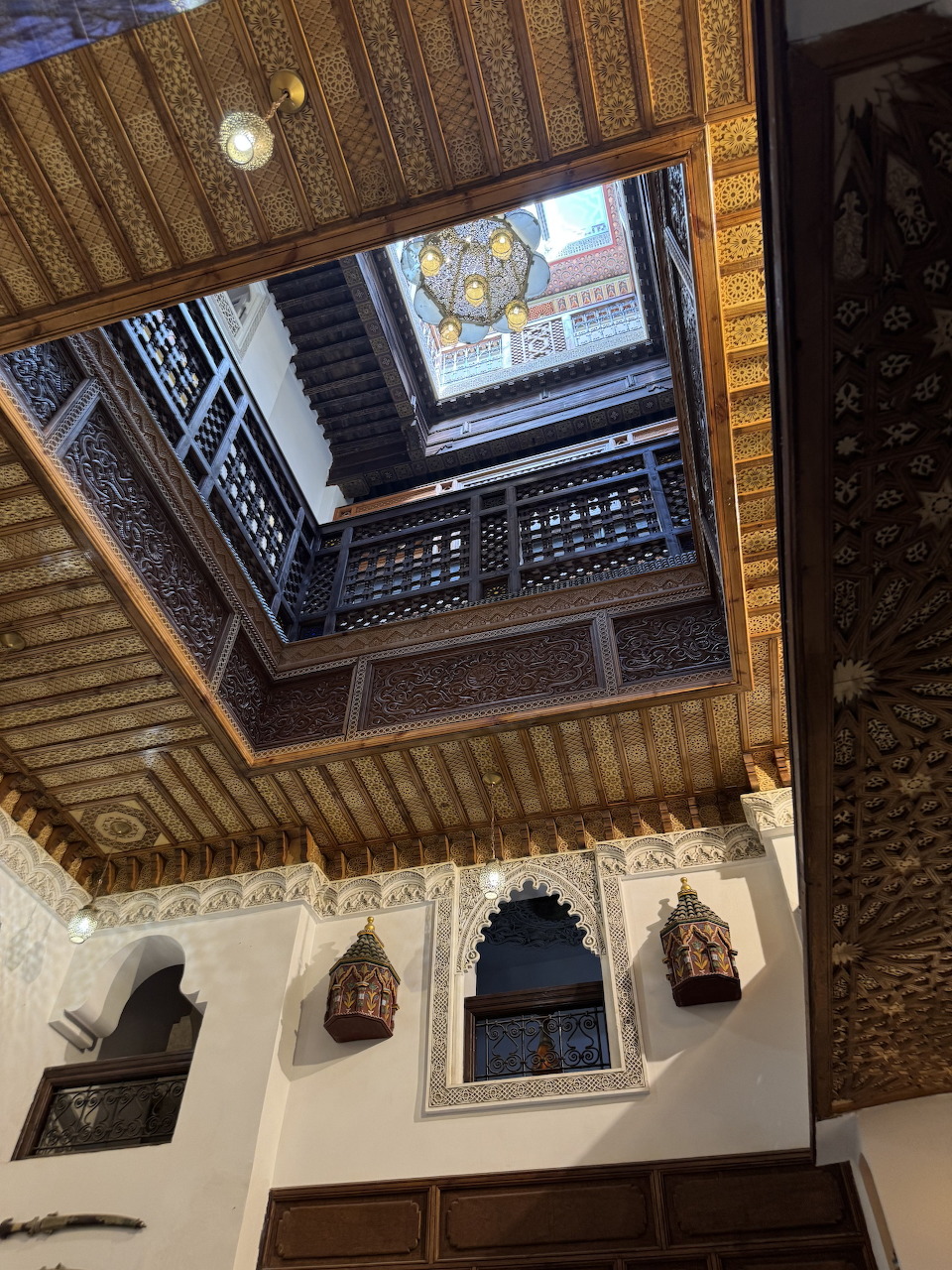 Upward view of ornate Moroccan riad courtyard with carved wood ceiling, white walls, and decorative arches