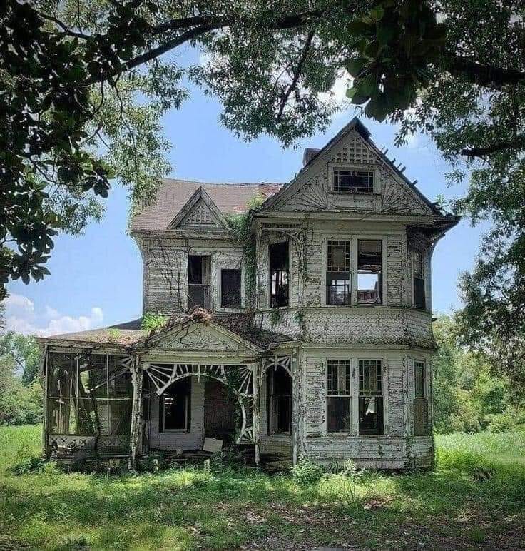 Deteriorating Victorian-era house with decorative porch trim surrounded by overgrown trees and grass