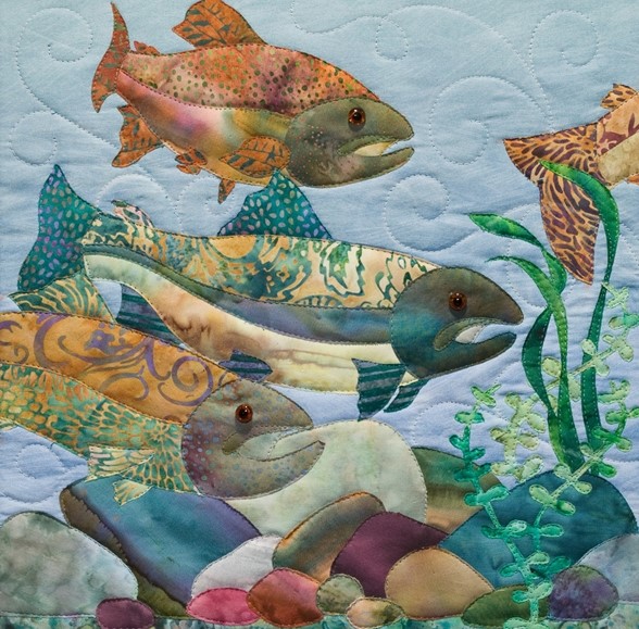 Quilted fabric art showing three patterned fish with aquatic plants and stones on textured blue background