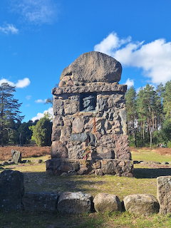 Large stone monument or cairn in a forest clearing with pine trees and blue sky