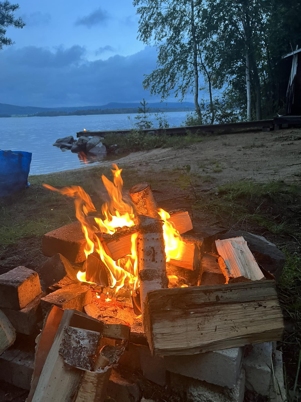 Campfire burning on a sandy lakeshore at dusk with trees and calm water in the background
