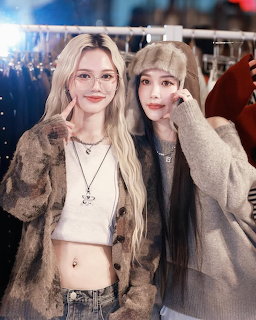 Two young women posing together indoors, one blonde in a crop top, one in a grey beanie