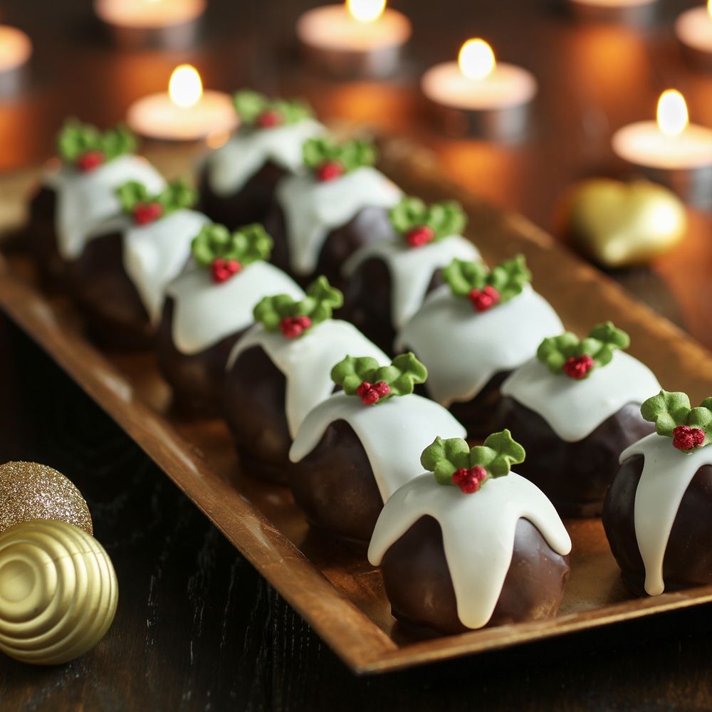 Mini chocolate Christmas pudding cakes with white icing and holly on a wooden tray