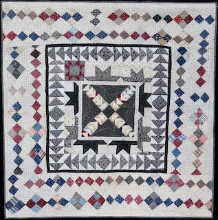 Detailed patchwork quilt with geometric patterns, diamond borders, and central star design in red, blue, and gray tones