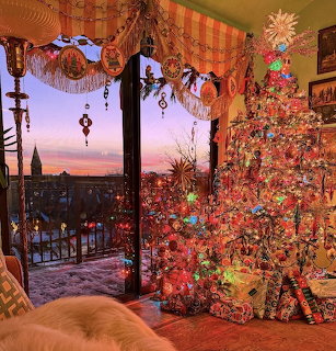 Illuminated Christmas tree with ornaments beside window showing sunset view over city rooftops