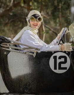 Woman in vintage white racing outfit and goggles driving a black antique race car with number 12