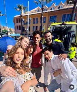Five young adults taking a selfie outdoors near a building and a yellow emergency vehicle