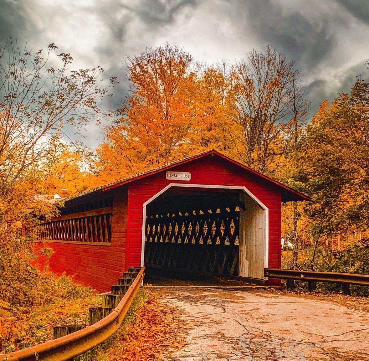 Red covered bridge framed by golden autumn trees beneath stormy gray clouds