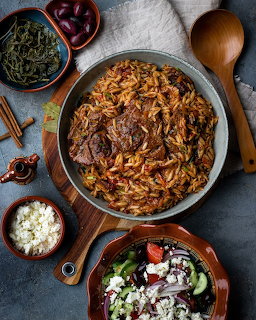 Overhead shot of Greek giouvetsi with slow-cooked beef and orzo in a pan, surrounded by Greek salad, feta, and olives.