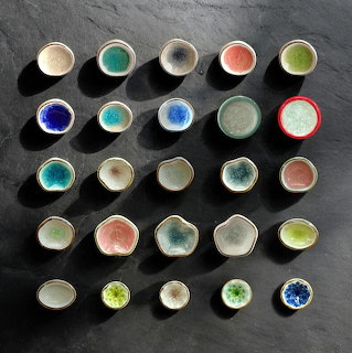 Twenty-five small ceramic bowls with colorful glazes arranged in rows on dark background